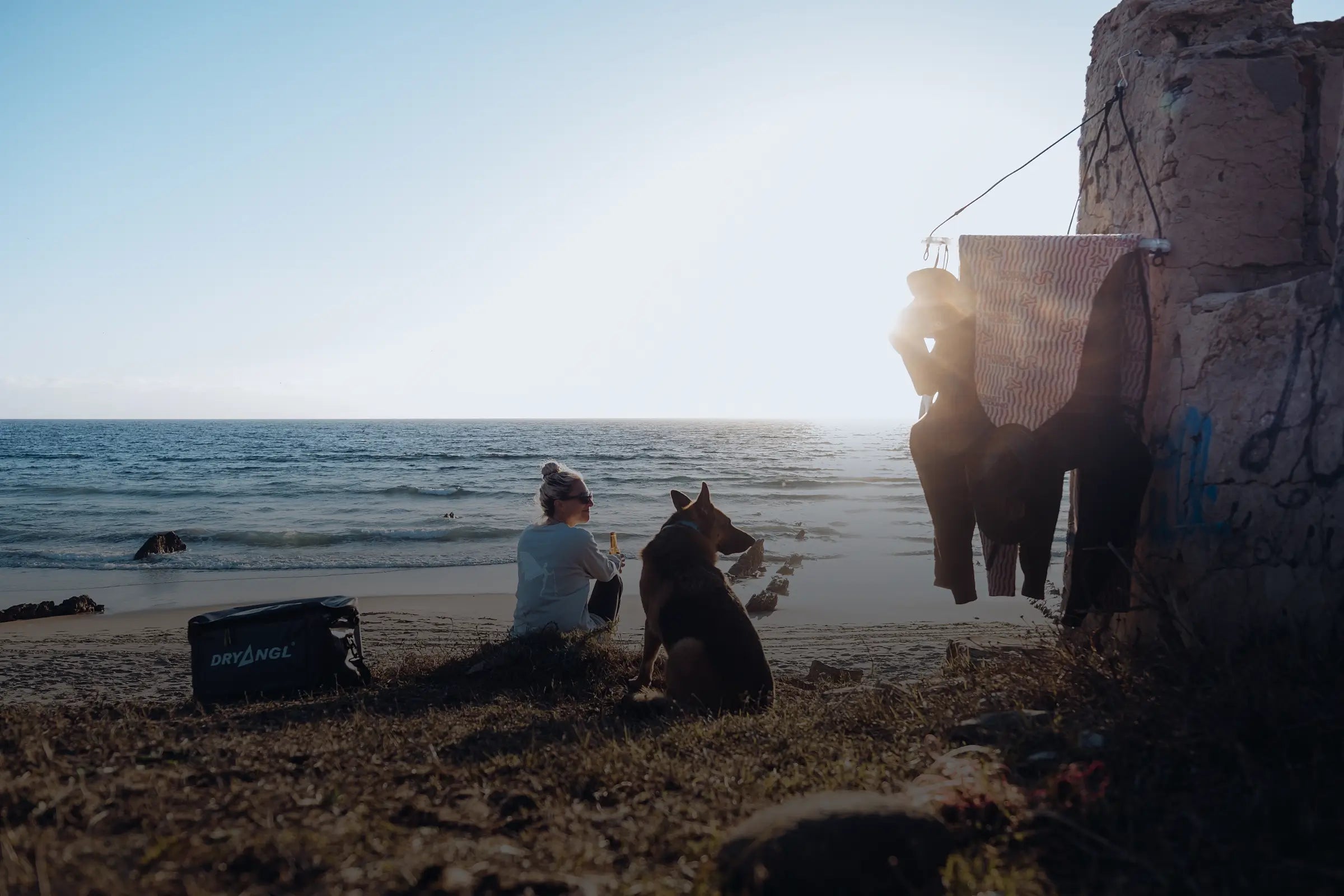 Frau mit Hund am Strand bei Sonnenuntergang und Dryangl Tasche im Vordergrund. Symbol für Freiheit und Outdoor-Lifestyle.